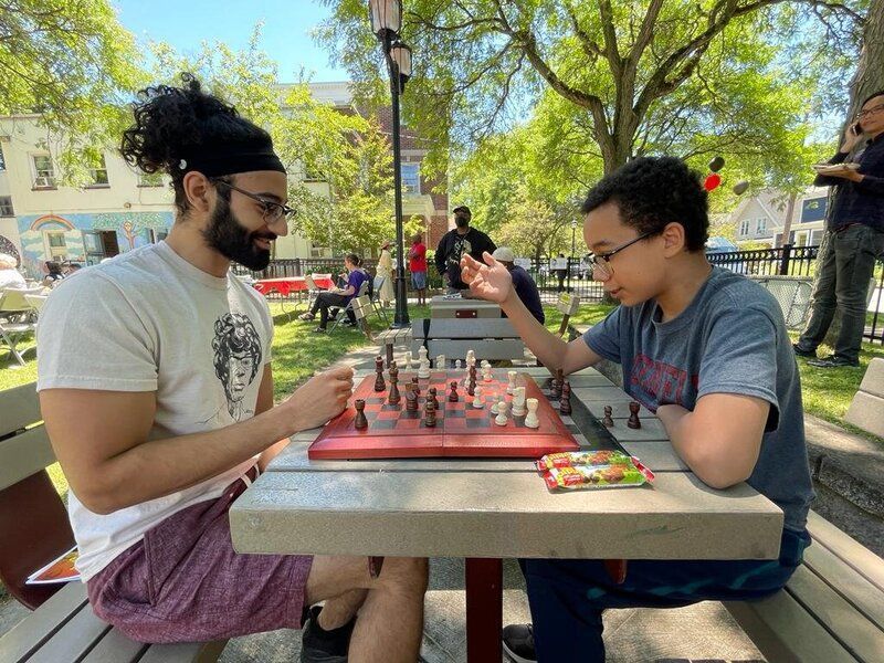 Two men are playing chess at a picnic table in a park.