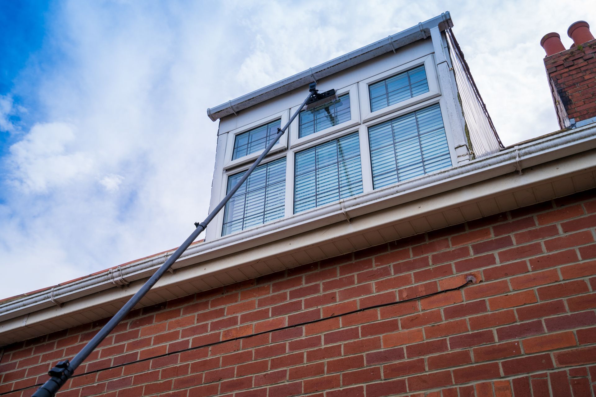 Window washer cleaning a window on a brick building with a long pole, against a cloudy sky.