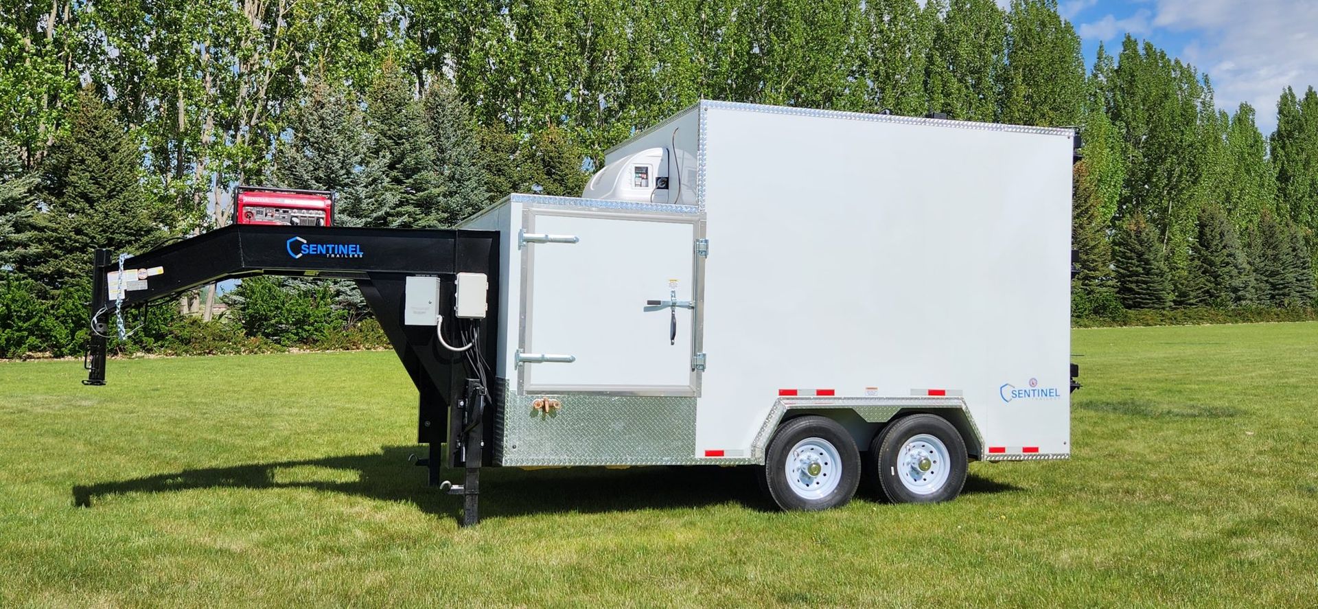 White enclosed trailer with dual axles parked on grass beside trees under a blue sky