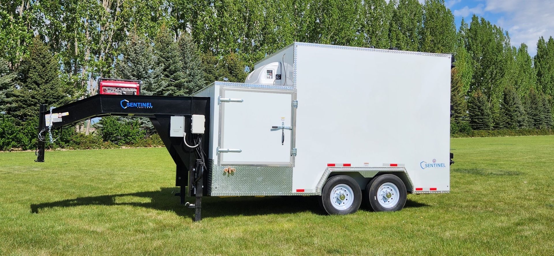 White enclosed meat processing trailer with black hitch and dual axles parked on grass by trees under a blue sky
