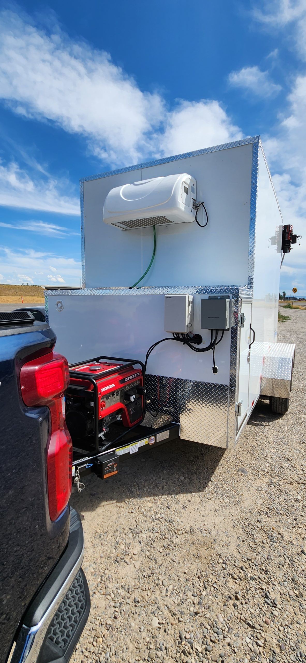 Portable generator mounted on a white butcher trailer beside a red pickup truck on a gravel lot under blue sky
