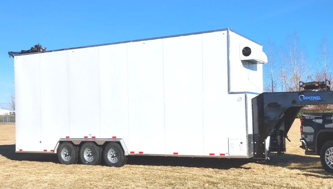 White enclosed butcher trailer with rooftop vent parked on gravel beside a pickup truck at sunset.