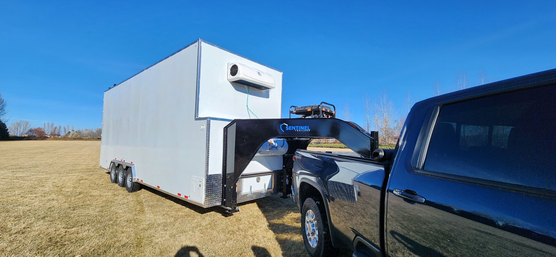 Black pickup truck parked beside a white box trailer in a grassy field under a blue sky.