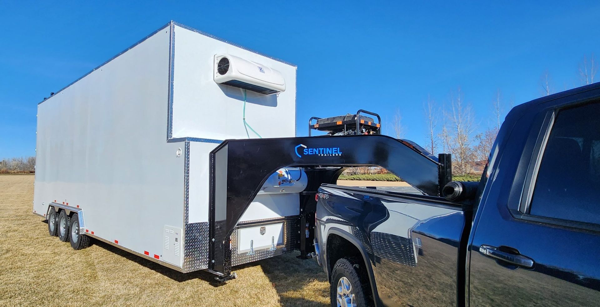 White enclosed Sentinel trailer with slide-out equipment attached to a pickup truck in a grassy field under a clear blue sky