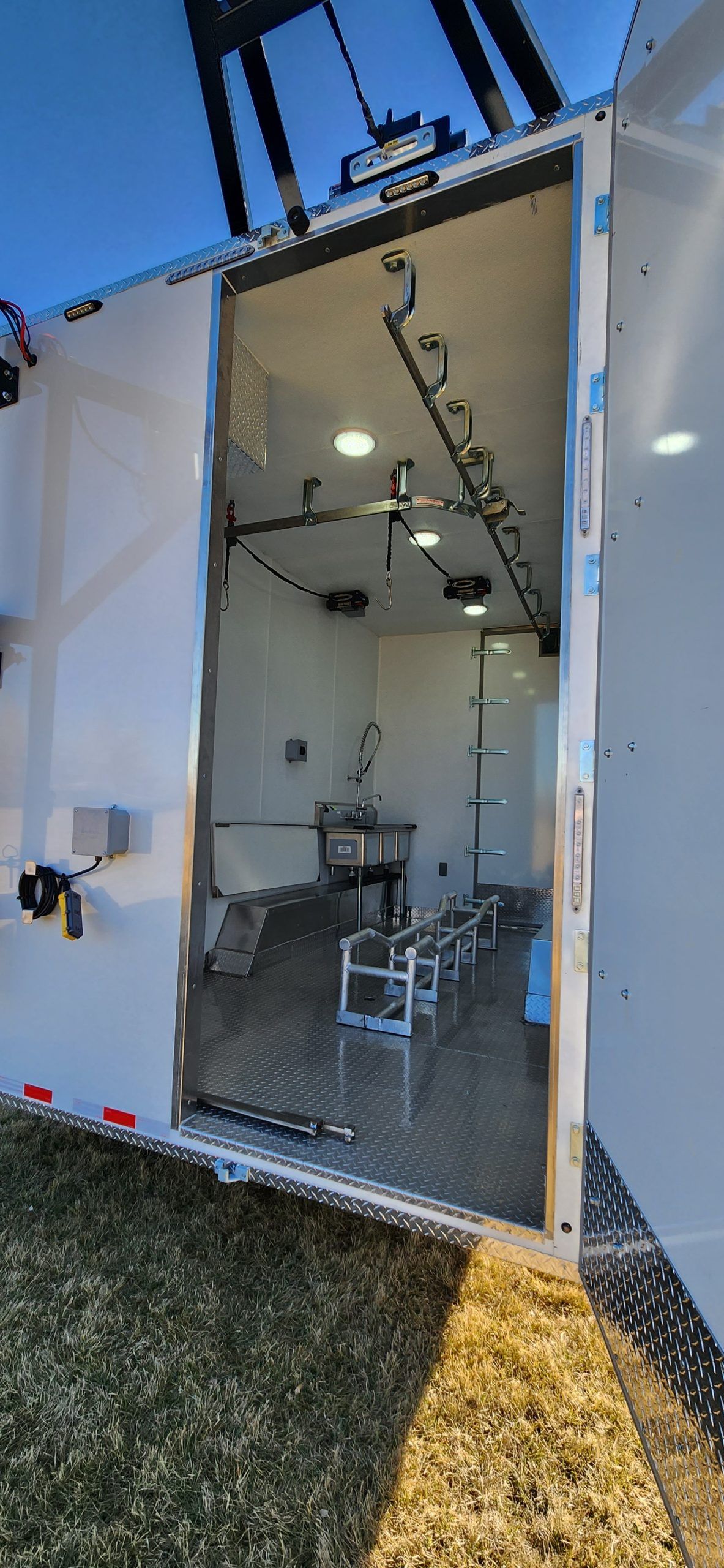 Open custom butcher trailer with a mirrored wall and overhead lights inside, parked on grass.