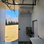 Interior of a butcher trailer with rails and open doorway to a sunny field outside.