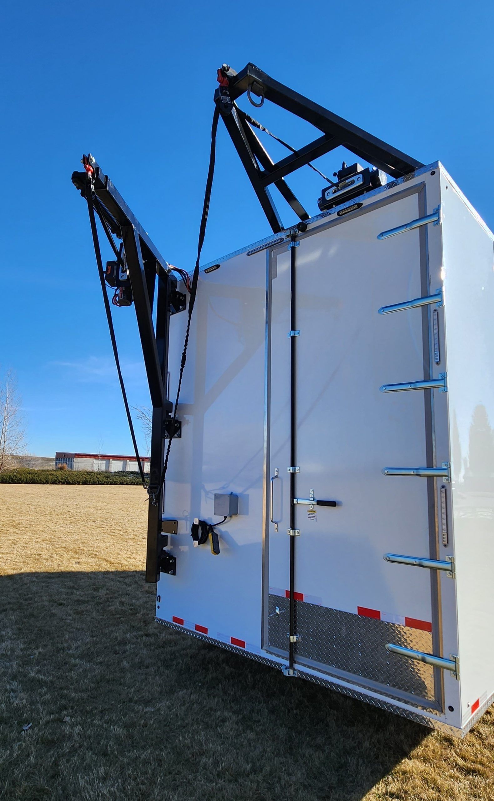 Rear view of a white utility trailer with raised black hydraulic arms in a grassy field under a blue sky