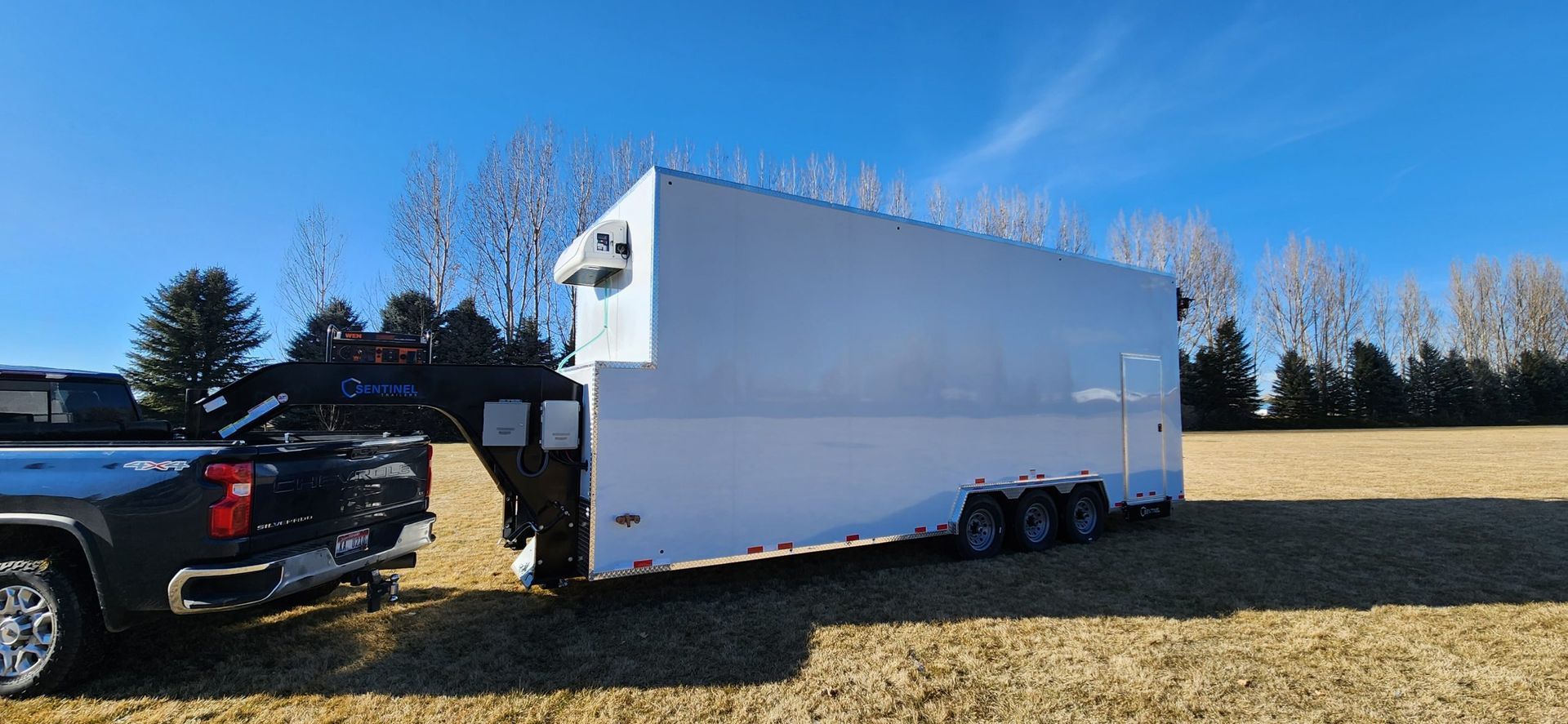 Black truck towing a large white enclosed meat processing trailer in a grassy field under a clear blue sky