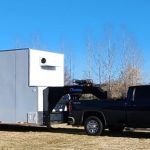 Black pickup truck parked beside a white trailer under a clear blue sky.