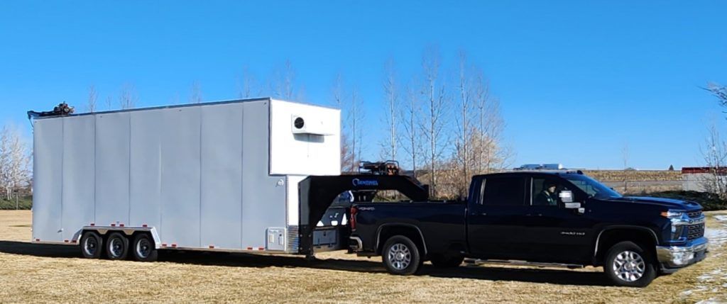 Black pickup truck towing a large white enclosed meat processing trailer on a grassy roadside under a clear blue sky