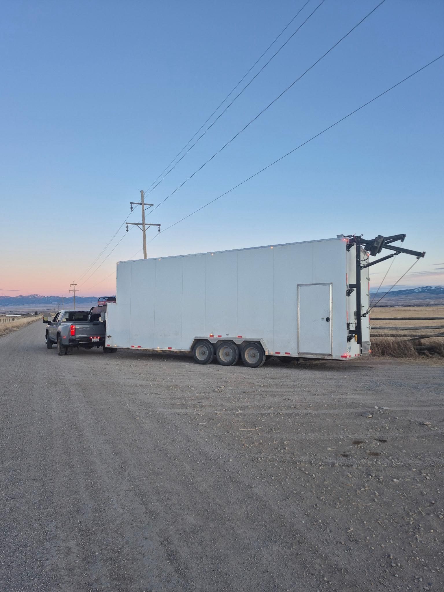 White enclosed trailer hitched to a pickup truck in a gravel lot at sunset.