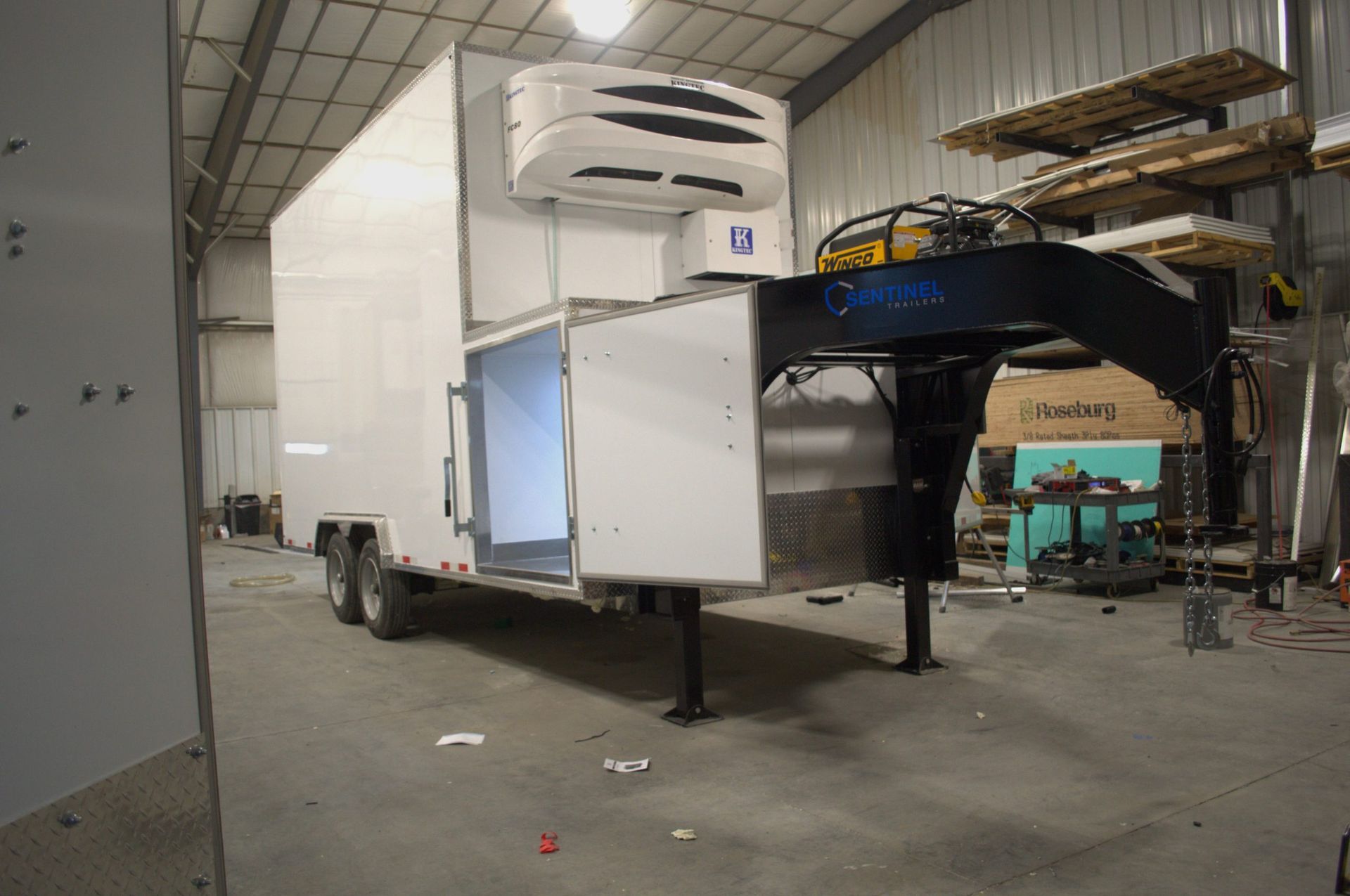 Large white meat processing trailer with black platform parked inside a warehouse, with shelves and equipment nearby