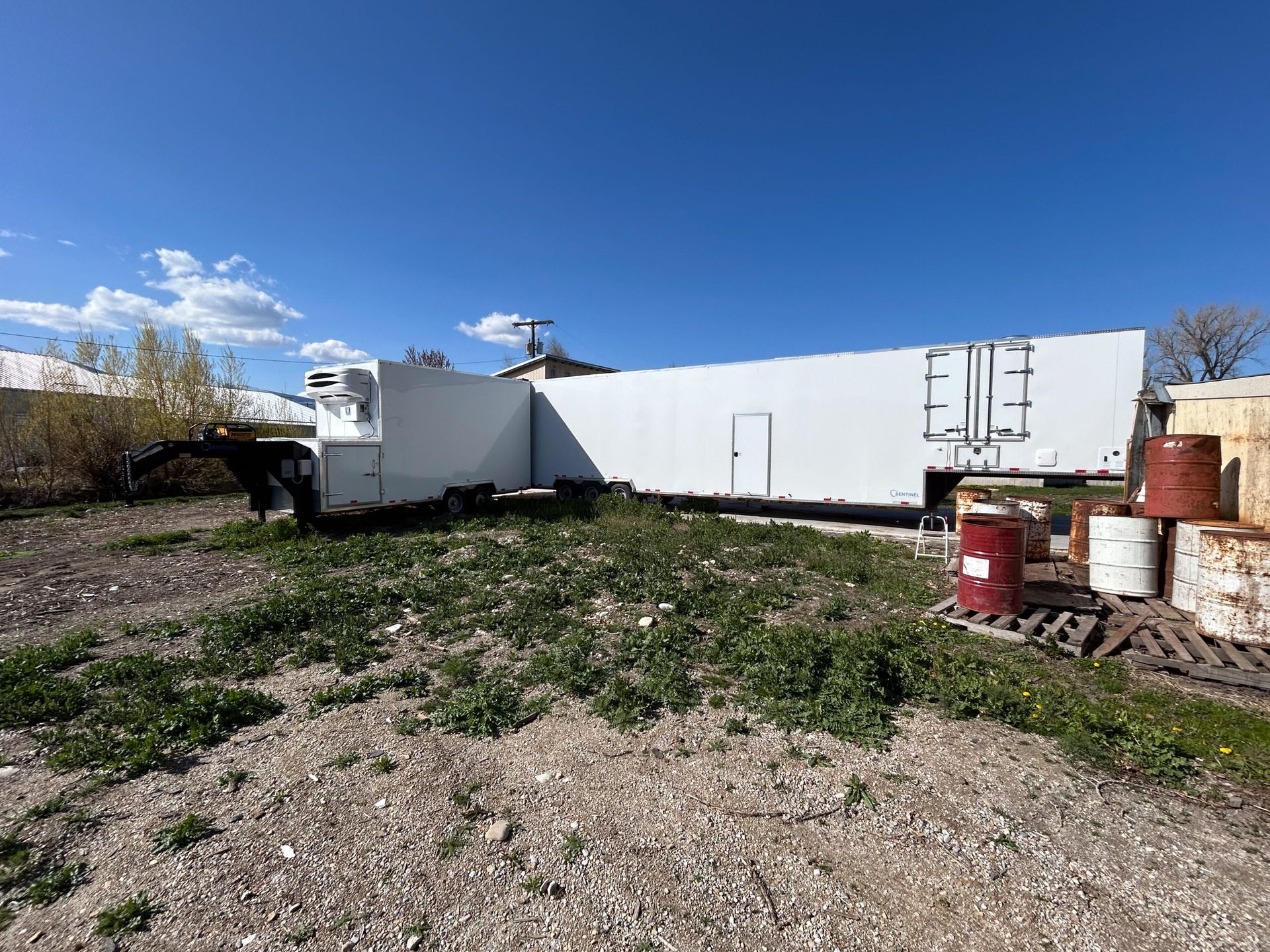 Open gravel lot beside a white building with red barrels under a blue sky