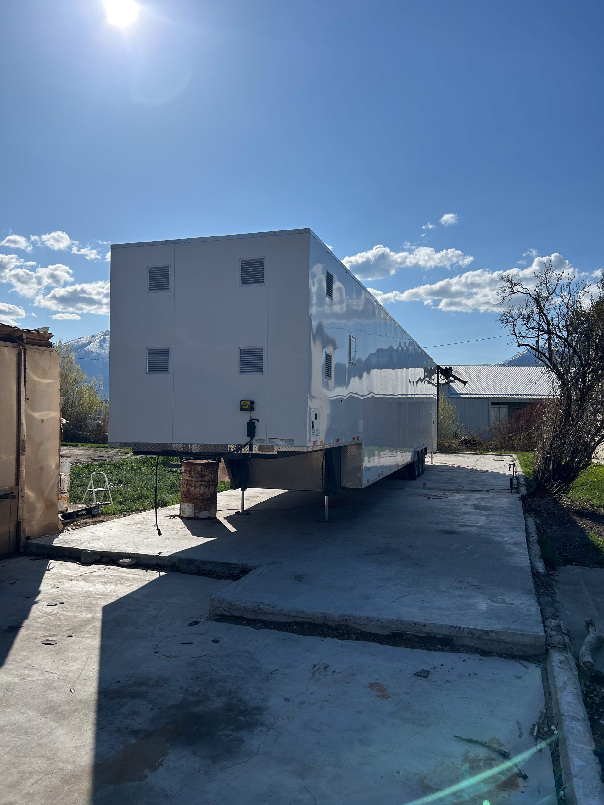 Large white trailer on a narrow paved road under a bright blue sky, near houses and greenery