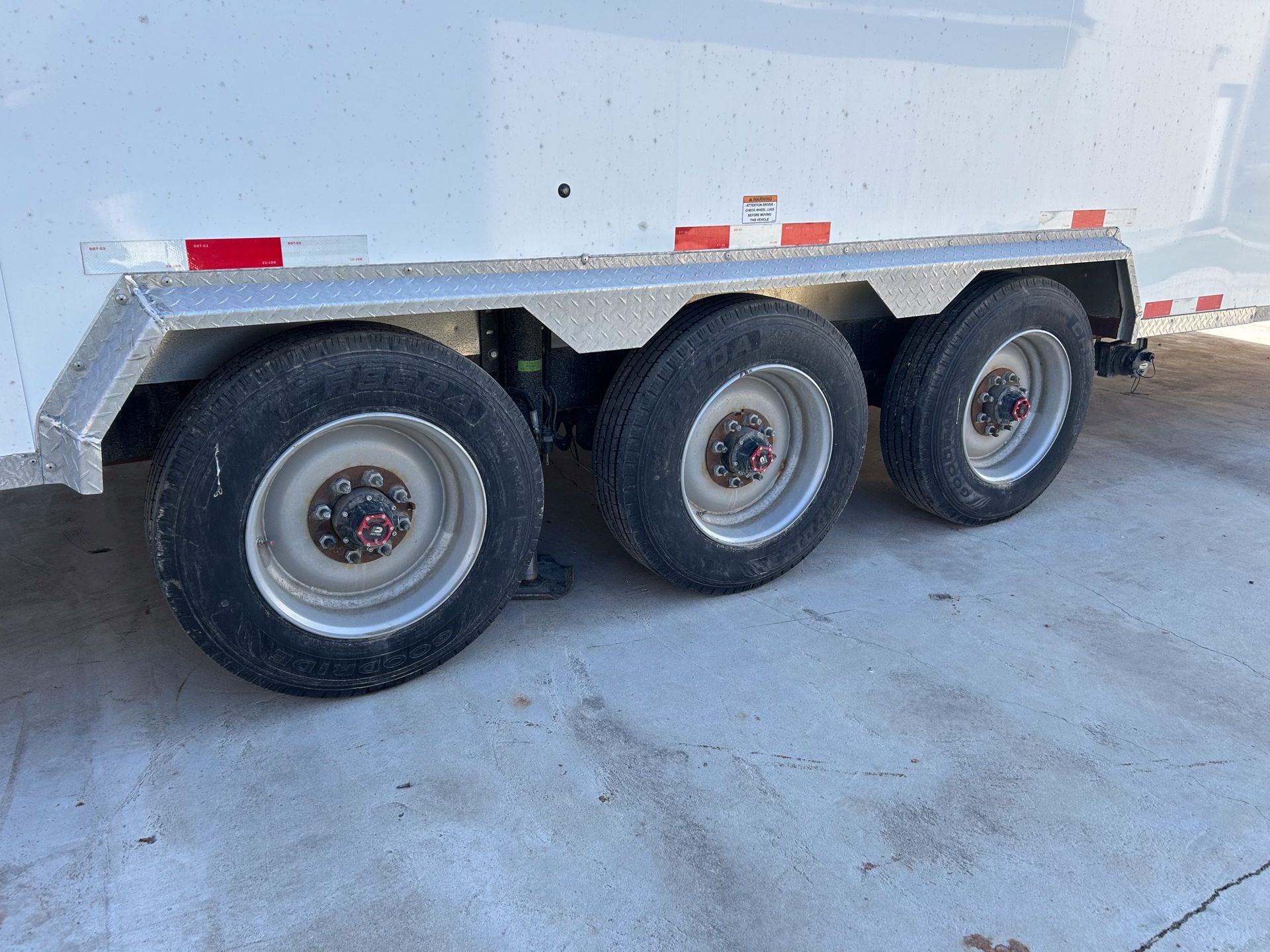 Three-axle utility trailer with silver sides and black tires parked on concrete near a wall