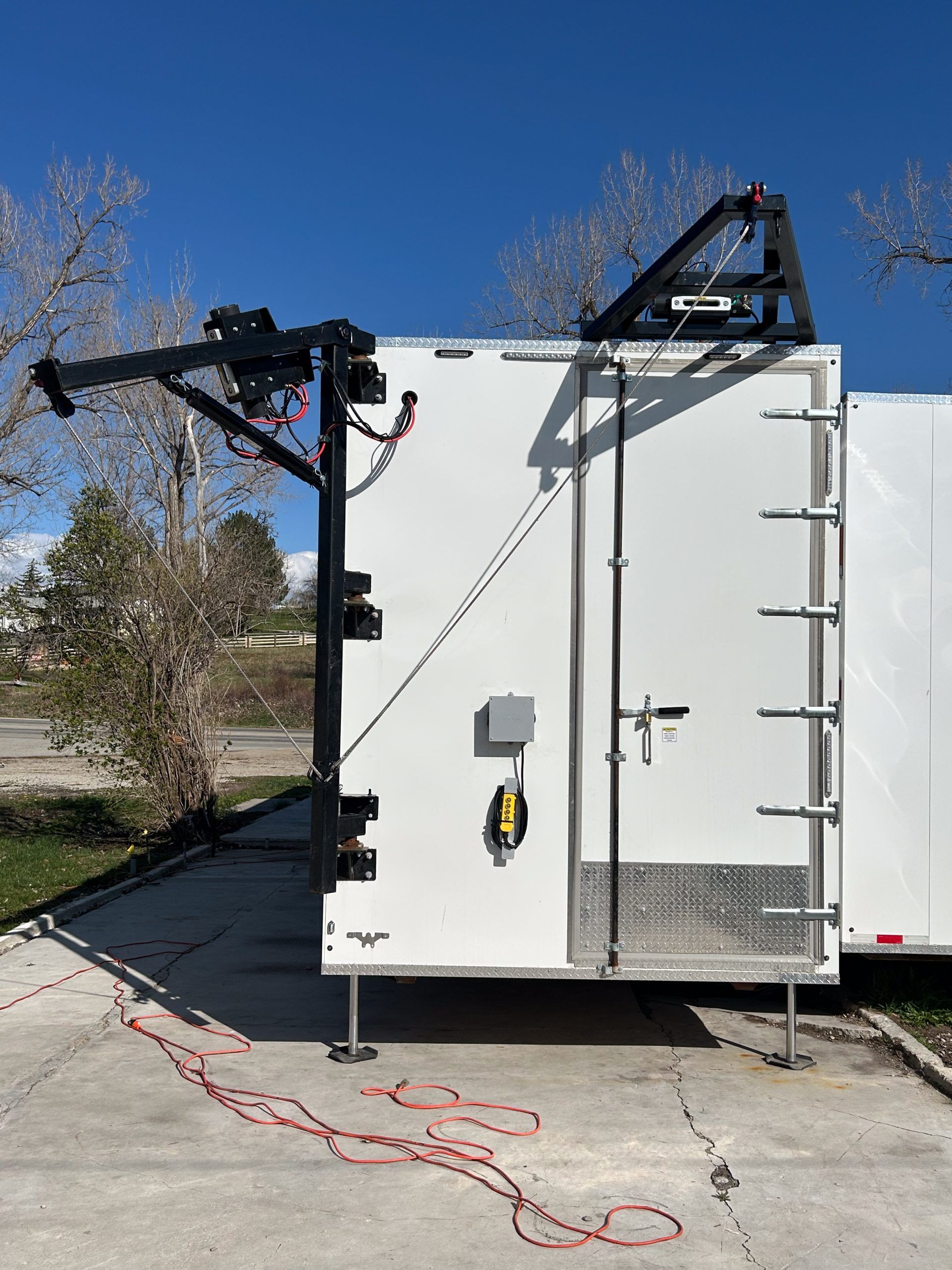 White utility trailer with mounted black equipment, parked outdoors on pavement under a clear blue sky.