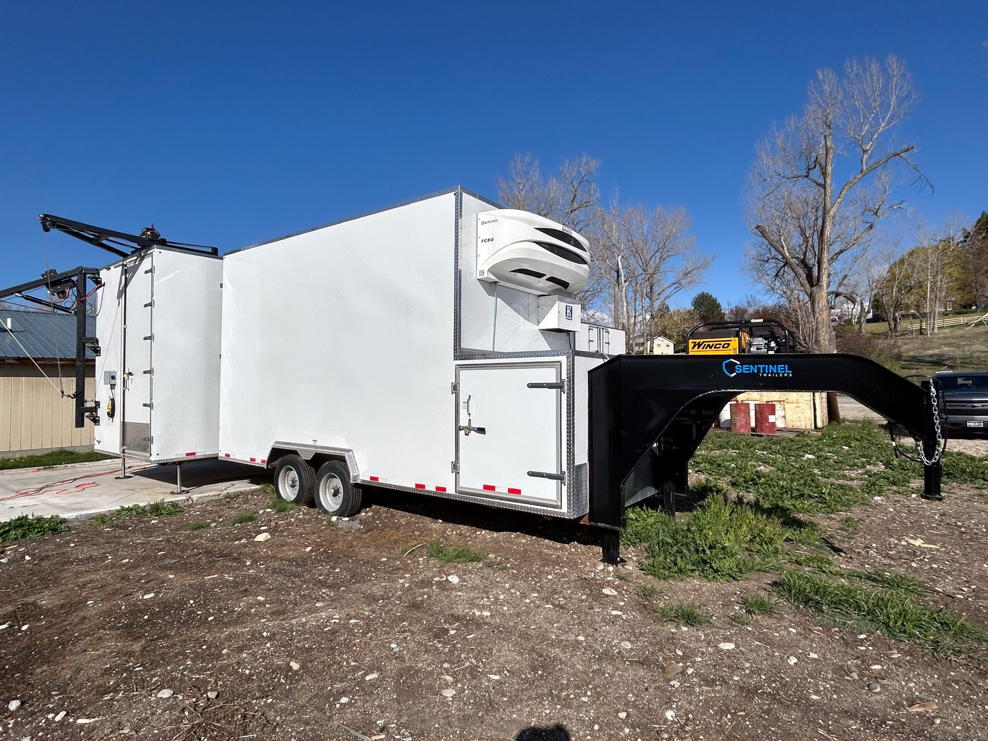 White enclosed trailer with an open black ramp parked on dirt near trees and a building.
