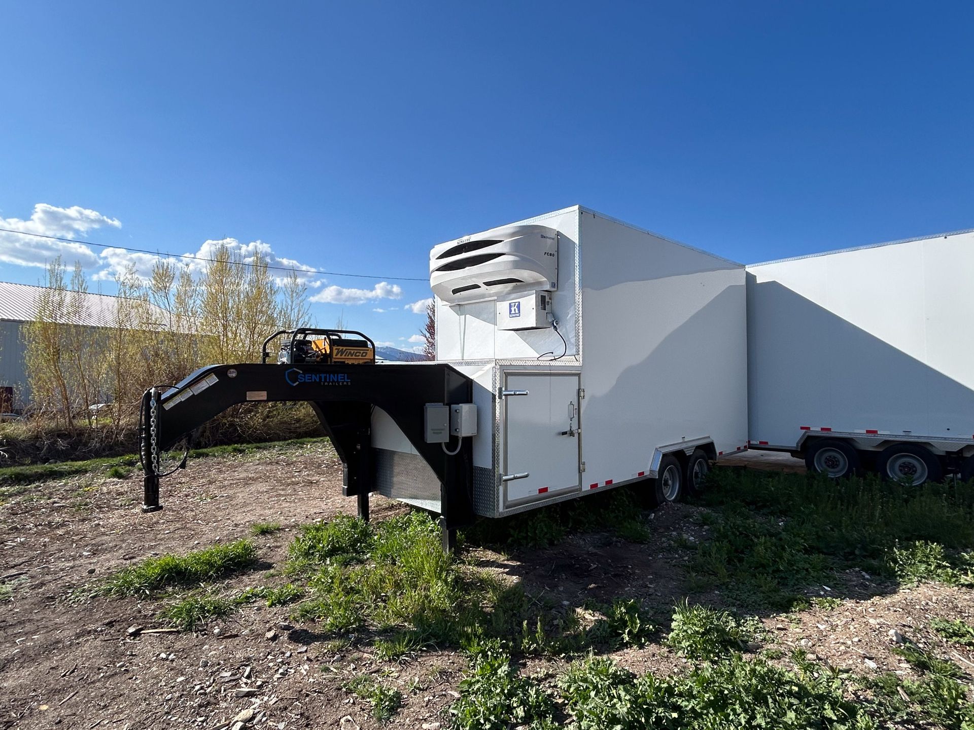 White box trailer with a black loading ramp parked on gravel under a blue sky