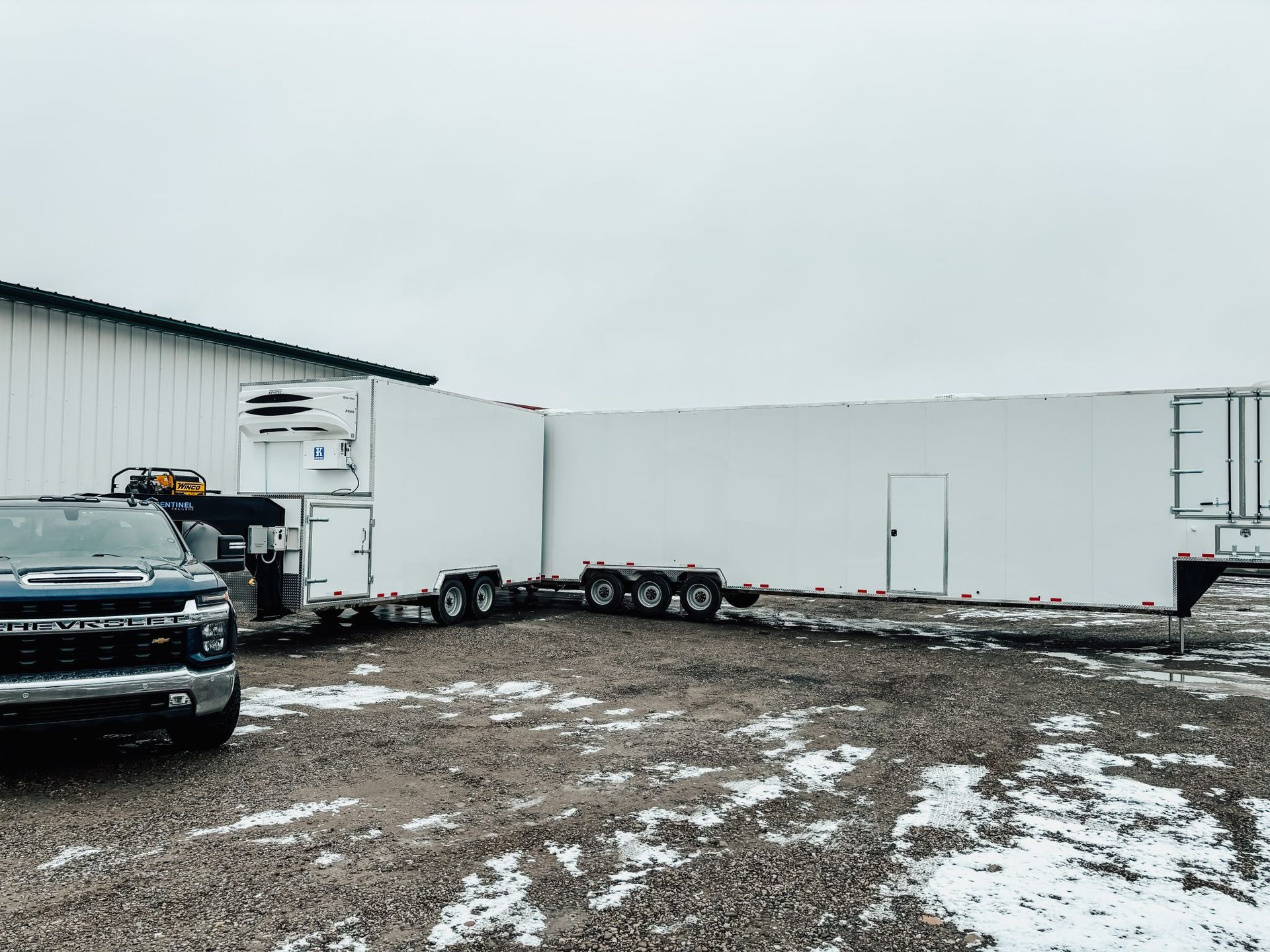 Snow-dusted industrial lot with semi-truck trailers for butcher beside a white warehouse and pickup truck