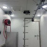White room with ceiling-mounted equipment, a red hose reel, and a wall ladder next to a door