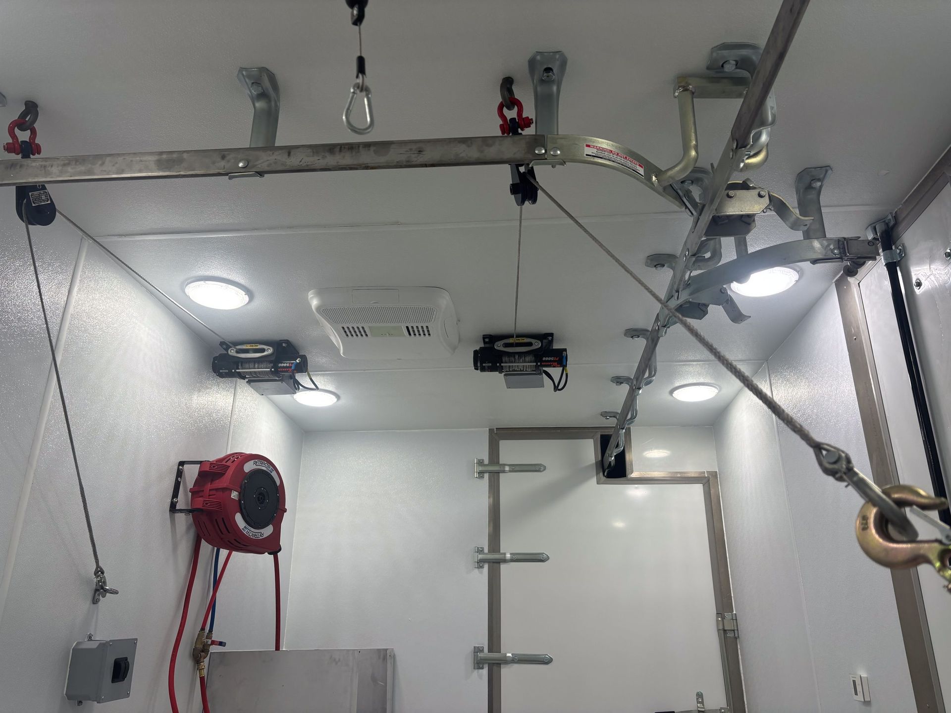 Empty white room with ceiling railings, hanging hardware, a red wall fan, and a ladder-like frame.