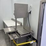 Metal machine on a wheeled cart in a custom made butcher trailer, with a white work table and cables nearby.