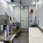 Industrial kitchen with stainless steel equipment, a worktable, and a white counter under bright lights in refrigerated