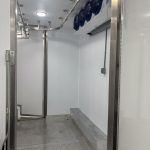 Empty industrial room with stainless steel pipes, white walls, and a gray tiled floor inside a mobile slaughter trailer
