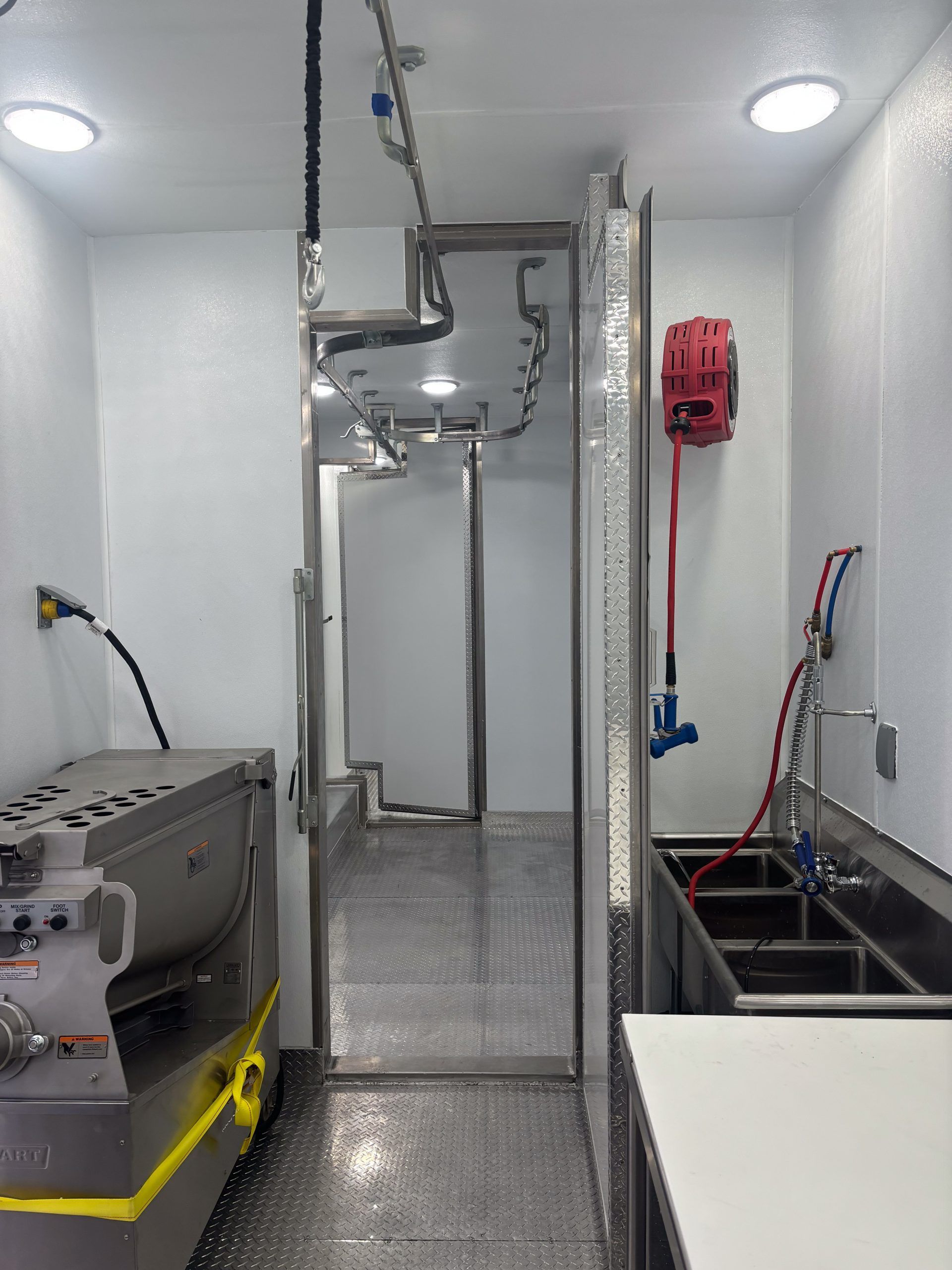 Narrow utility room with metal equipment, hose reels, and a central mesh gate under bright ceiling lights