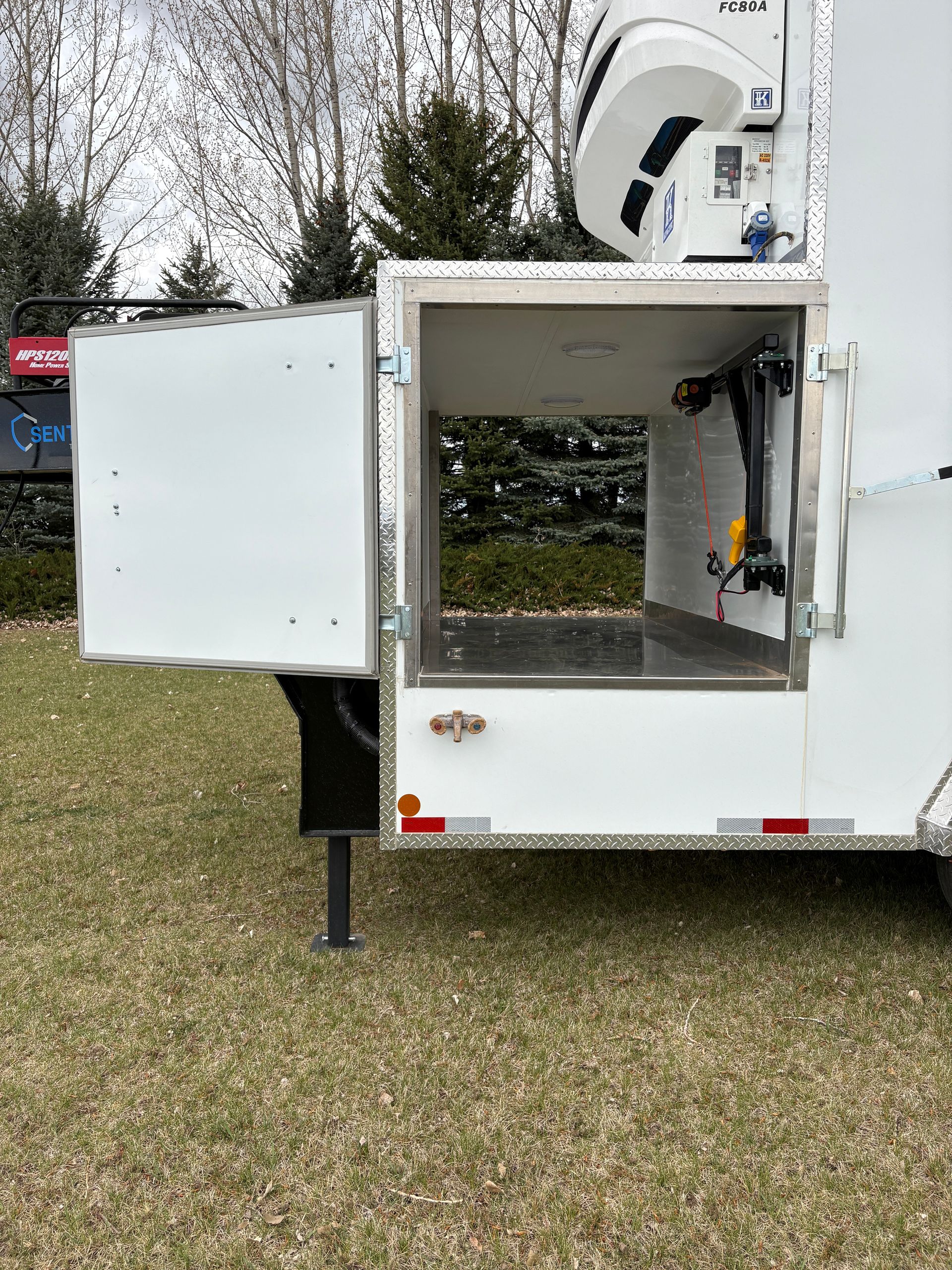 White utility trailer with open side door and raised antenna, parked on grass outdoors.