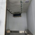 Empty metal utility shaft with white walls, overhead pipes, and a small blue vent near the ceiling.
