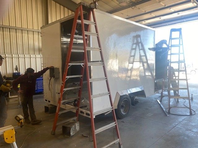 People assembling a white cargo mobile slaughter trailer in a workshop, with ladders leaning against it. 