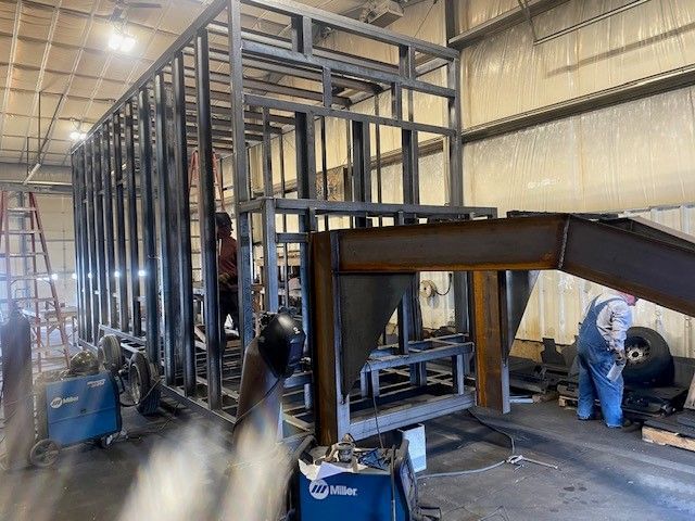 Workers welding a steel frame for a mobile meat processing trailer inside an industrial workshop
