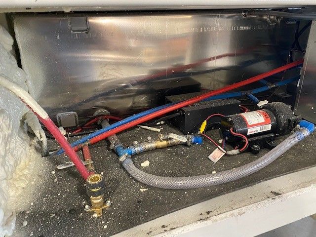 Under-sink plumbing with red and blue water lines, hoses, and a pump assembly in a metal compartment