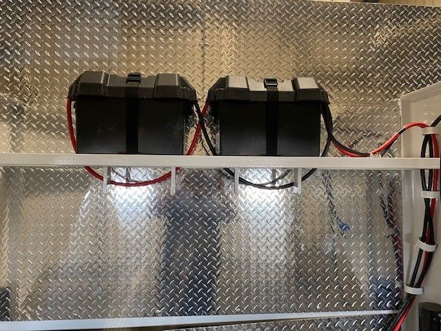 Two black battery packs mounted on a metal wall panel, with red and black wires and two small lights below.