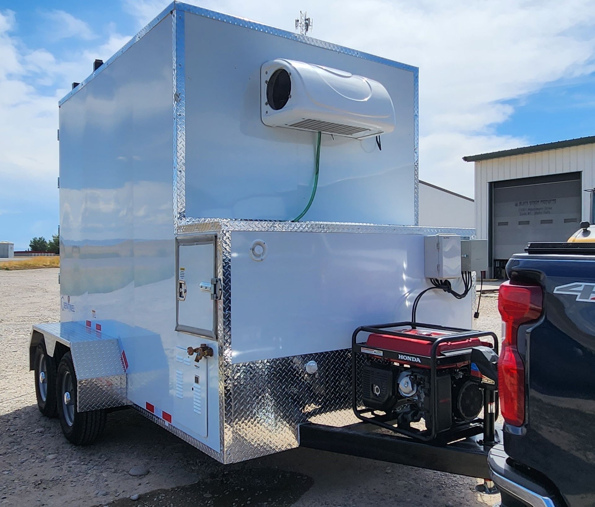 Silver mobile butcher trailer with a wall-mounted air conditioner and generator parked outdoors near a truck