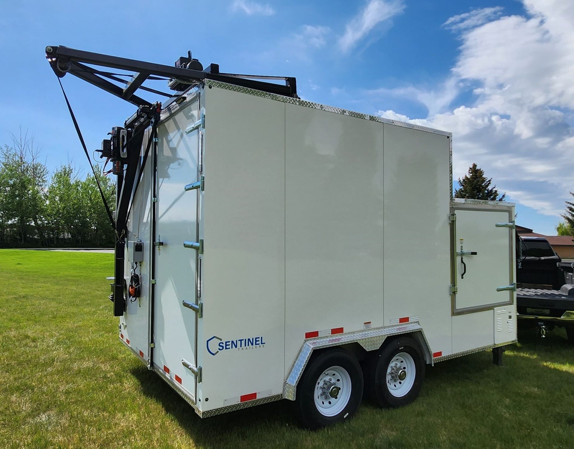 White utility trailer with a black hydraulic arm parked on grass under a blue sky.