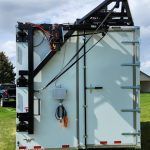White industrial trailer with black roof-mounted machinery and hoses parked on grass outdoors