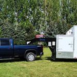 Blue pickup truck towing a white enclosed refrigerated trailer on grass with trees in the background