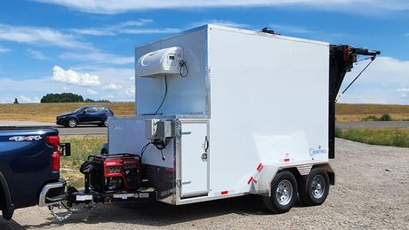White enclosed refrigerated trailer with open side compartment and generator parked beside a black truck on a gravel lot.