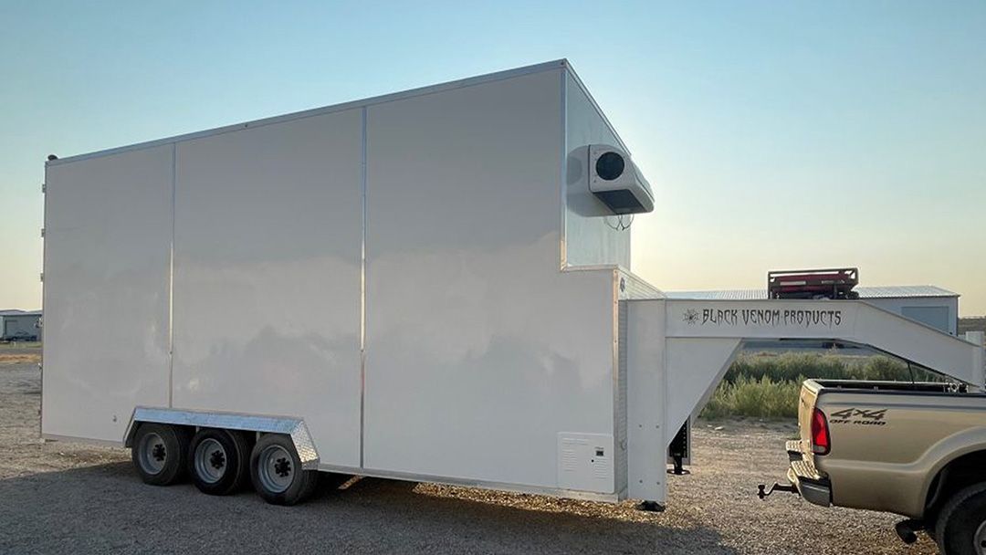 White enclosed trailer with rooftop vent parked on gravel beside a pickup truck at sunset.