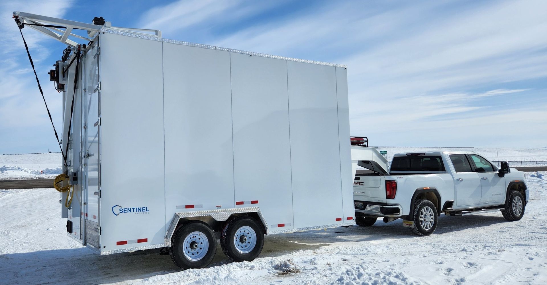 White pickup truck towing a large white enclosed trailer made by SEntinel trailers across a snowy landscape under a blue sky