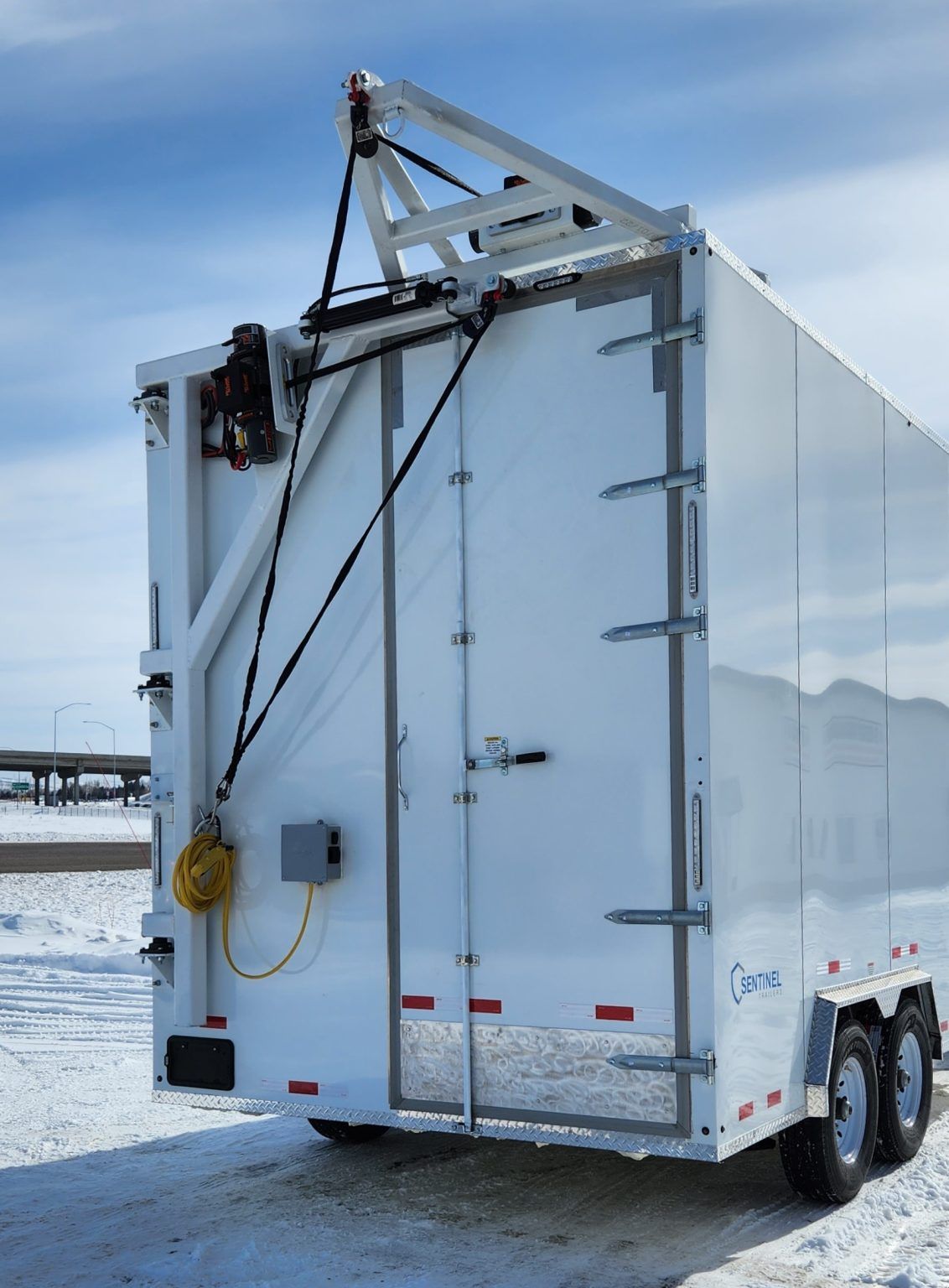White utility trailer with roof-mounted crane on a snowy roadside under a blue sky
