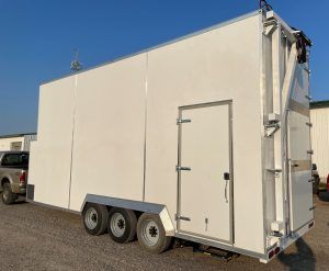 White enclosed trailer with three axles parked outdoors on gravel under a clear sky.