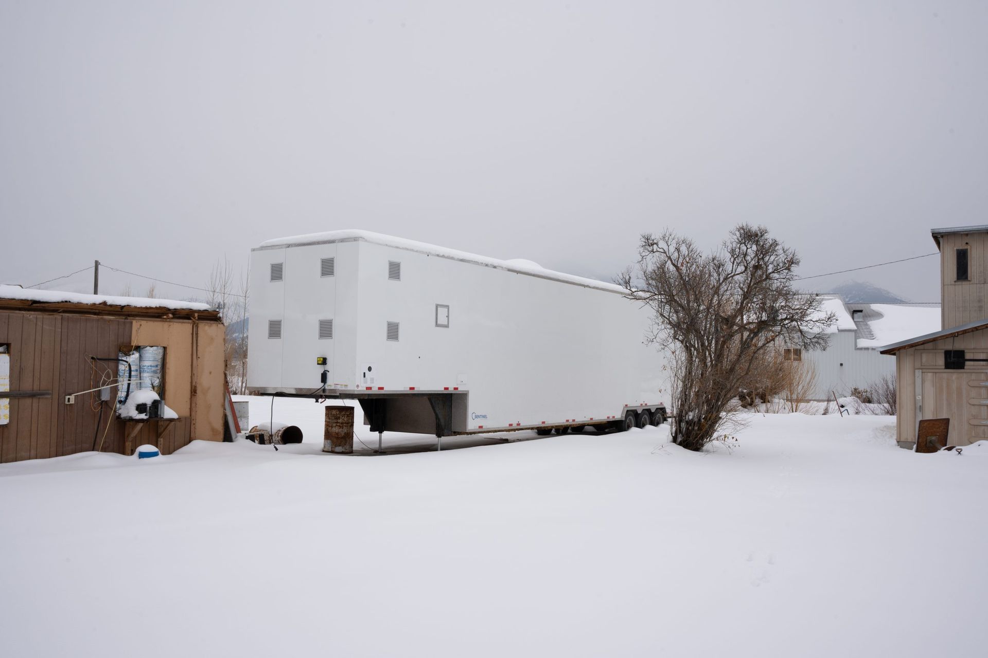 Snowy rural yard with white buildings, a bare tree, and overcast sky