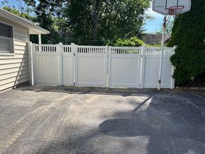 A white fence is surrounding a driveway next to a basketball hoop.
