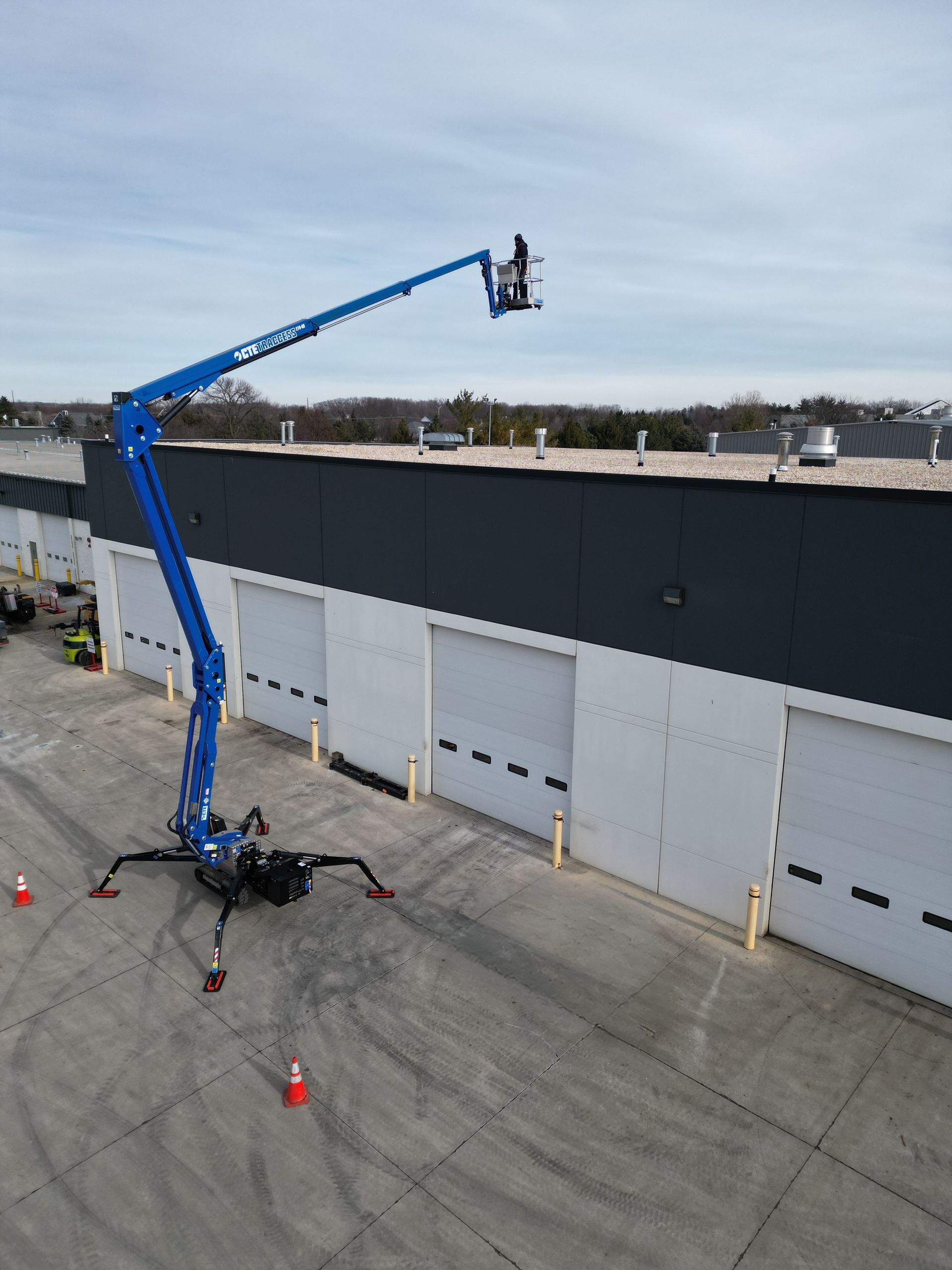 Blue boom lift reaching towards a building's roof; person in bucket. Orange cones on the ground.