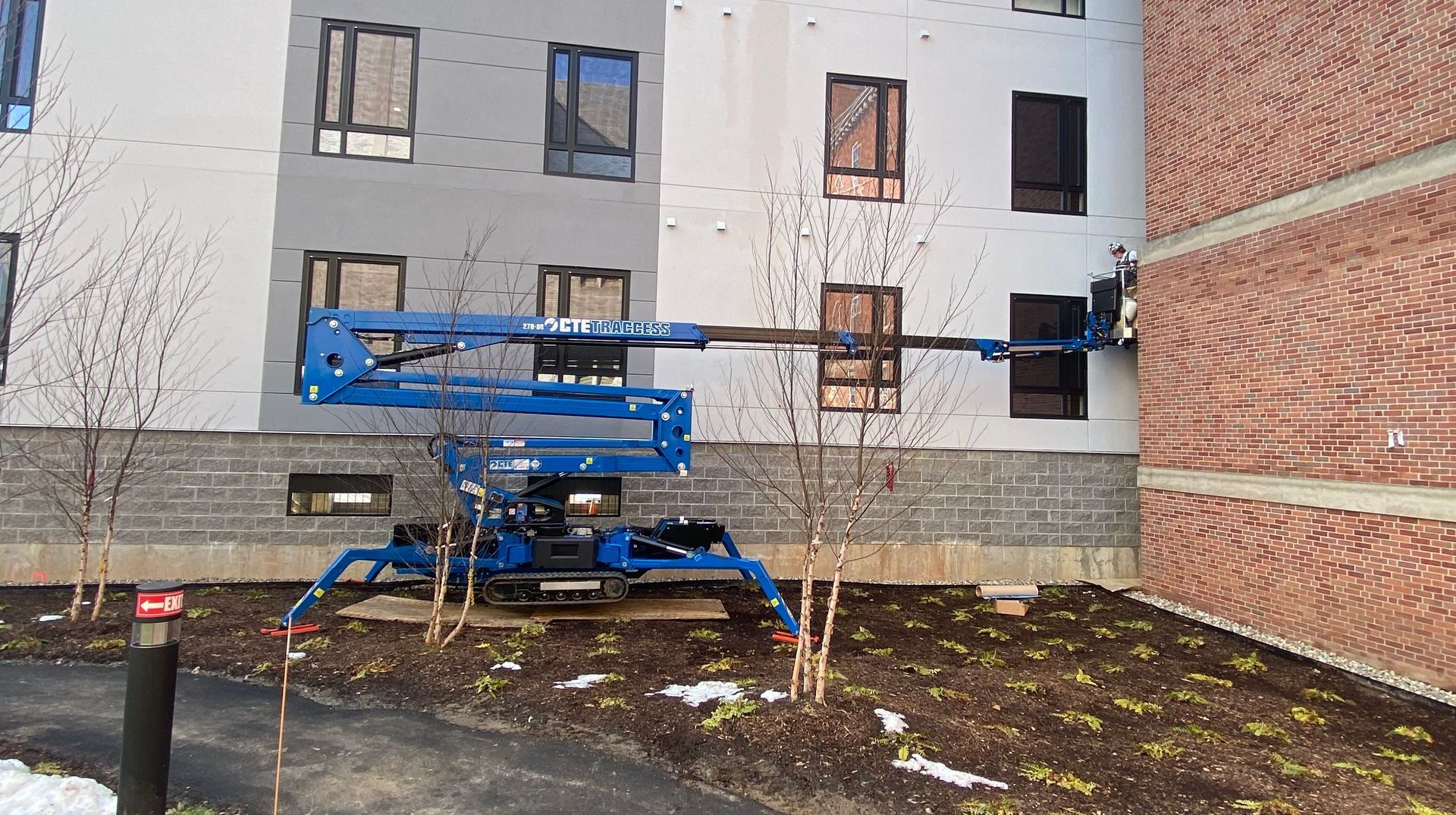 Blue lift reaching building's exterior. Construction worker at work, gray/white wall next to brick.