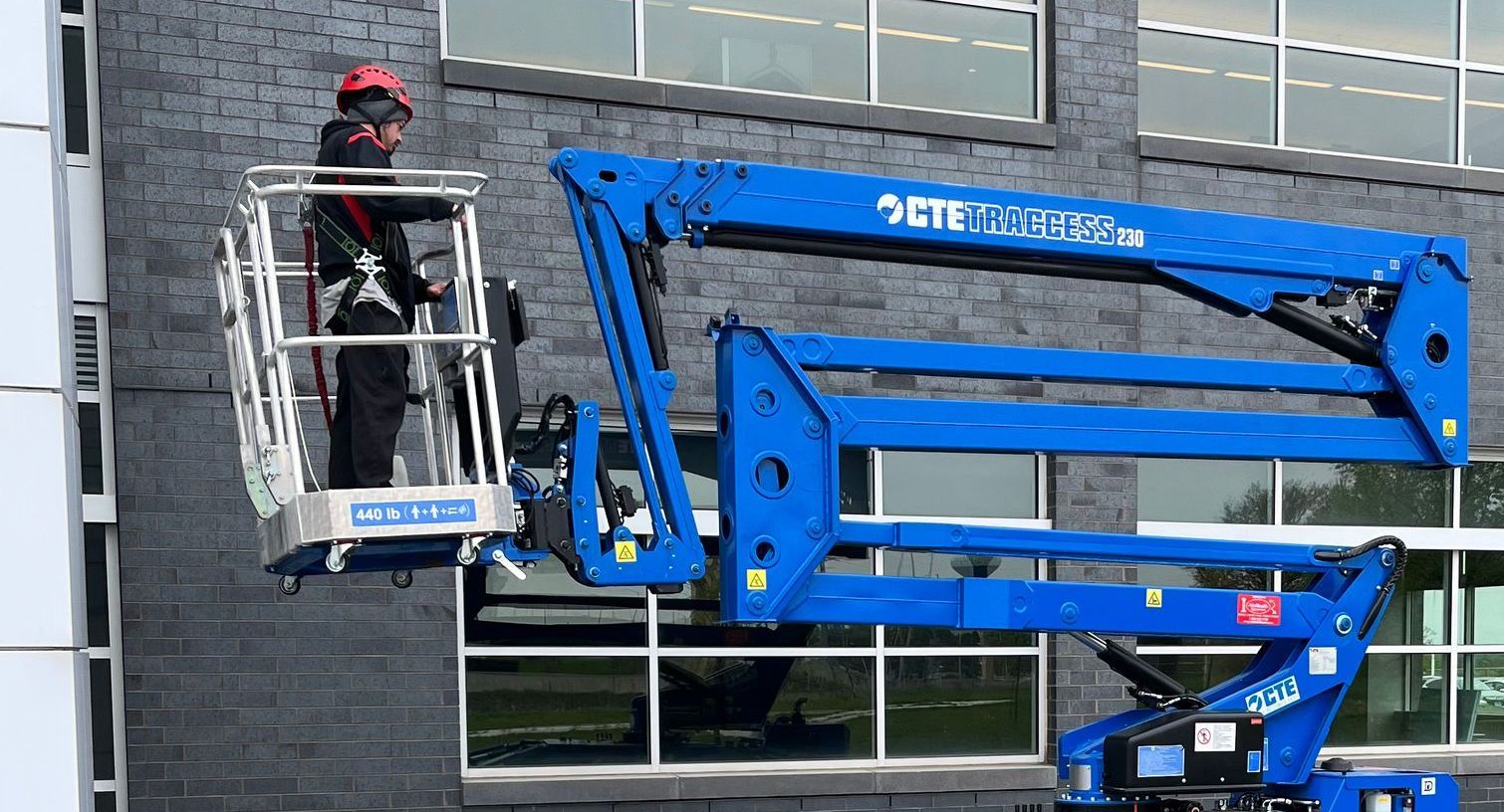 Person in an aerial lift near a building, wearing a helmet, working. Blue lift, gray brick wall, sunny.