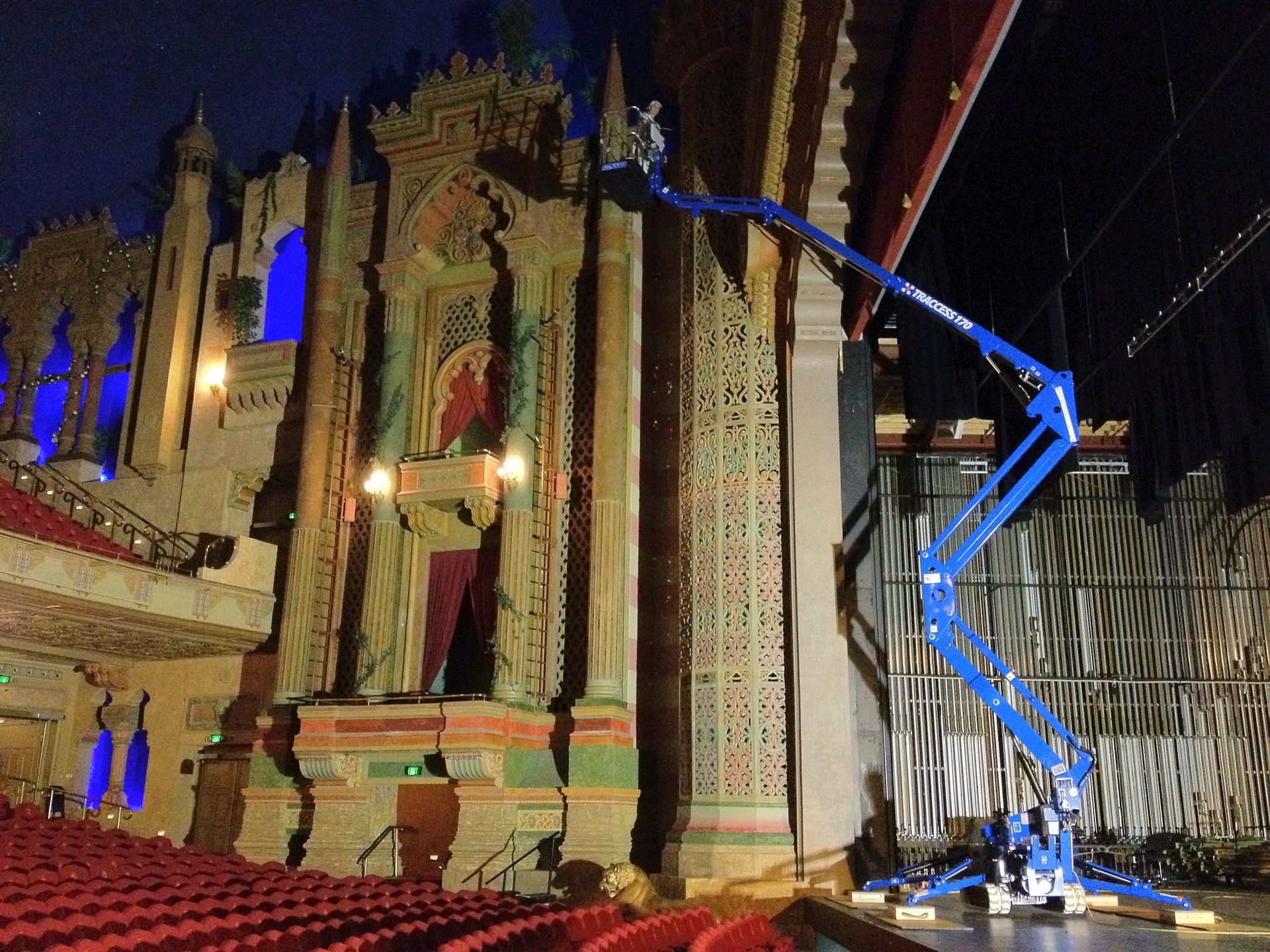 A worker in a blue lift adjusts ornate architecture in a theater. Red seats and stage lights visible.